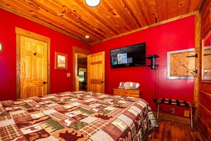 Une pièce avec un plafond en bois, des murs rouges, un lit recouvert d’une courtepointe à motifs, un téléviseur fixé au mur et une armoire en bois.