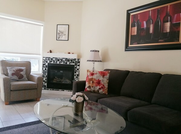 Una sala de estar con chimenea, un sofá, una mesa de centro de vidrio y un sillón con un cojín de un alce.