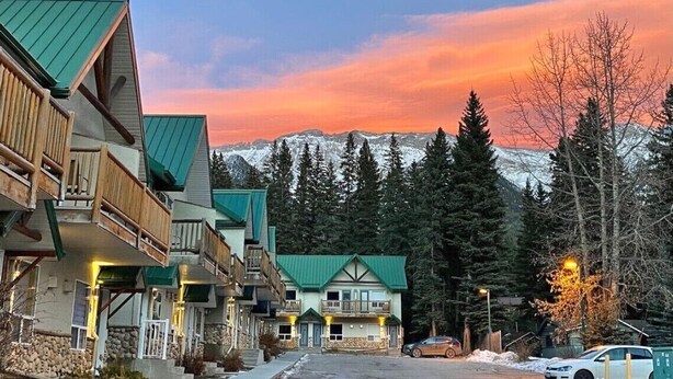 A row of wooden buildings with balconies, a central building with a green roof, and a mountain backdrop. - Promo Holidays!2Beds2Bath6PPL Banff Close Gem in Mountain View Townhouse (Harvie Heights)