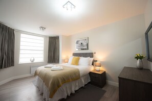 A modern hotel room with a neatly made bed, a bedside table with a lamp, a large window with curtains, and a framed picture on the wall.