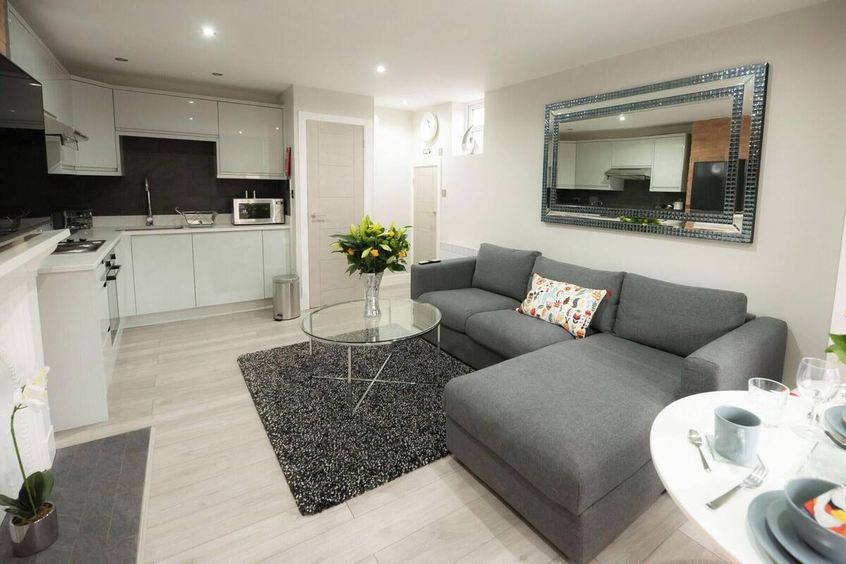 A modern living room with a grey sofa, a glass coffee table, and a dining area with a white table set for two.