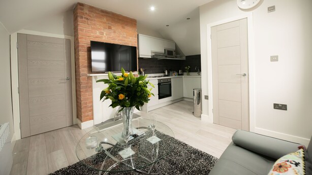 A modern kitchen with a brick accent wall, a dining area with a glass table, and a gray sofa. - Laurel House (Reading)