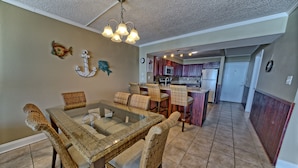 A dining area with wicker chairs and a glass-top table, a kitchen with wooden cabinets and stainless steel appliances, and a hallway leading to a door. - Oceanfront Dunes B406 + Free Seasonal Beach Loungers (Panama City)