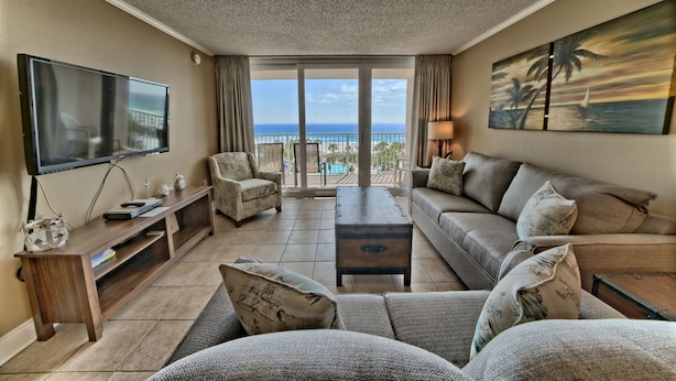 A spacious living room with a large flat-screen TV, comfortable seating, a wooden coffee table, and a view of the ocean through large sliding glass doors. - Oceanfront Dunes B406 + Free Seasonal Beach Loungers (Panama City)