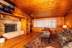 A cozy log cabin interior with a stone fireplace, wooden walls, a large window, and a television mounted above the fireplace.