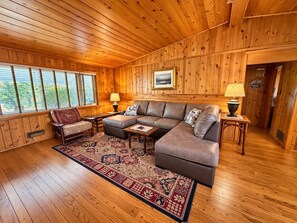 A cozy room with a wooden ceiling and walls, a sectional sofa, a small table, a chair, and a patterned rug.