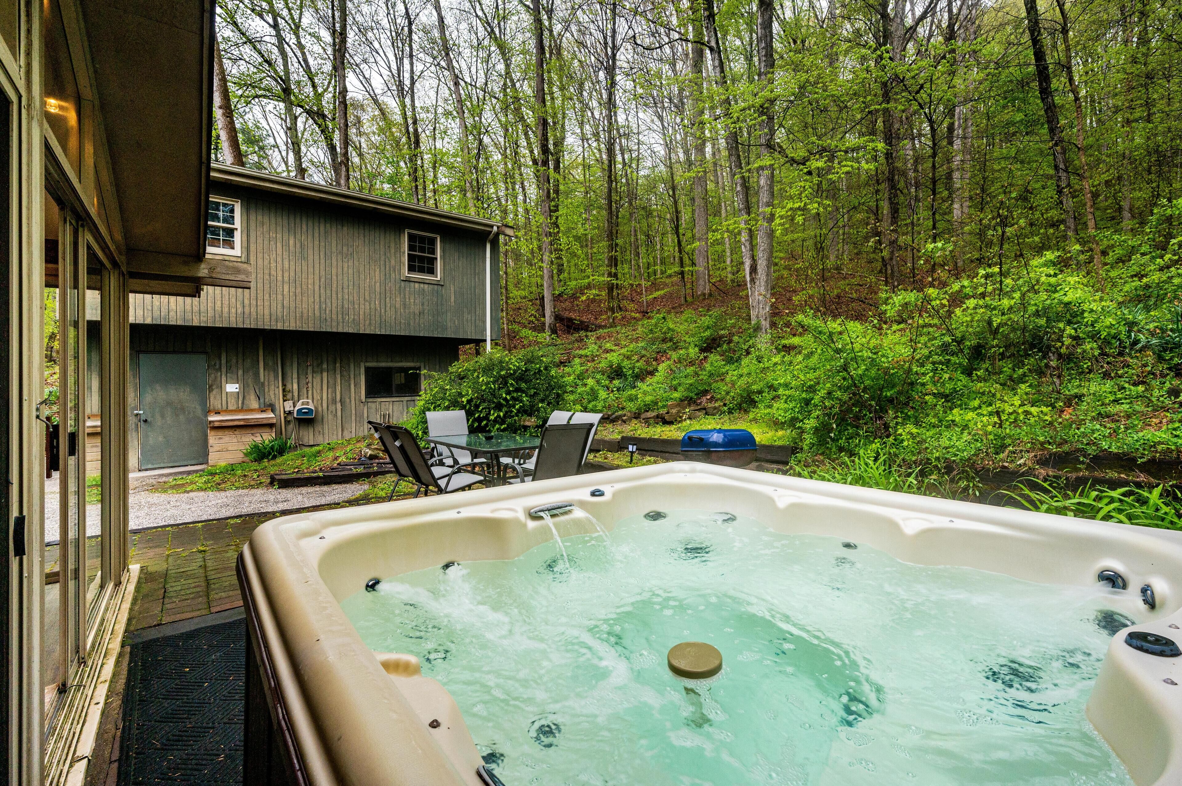 A hot tub in a forested area with a wooden cabin and outdoor furniture.