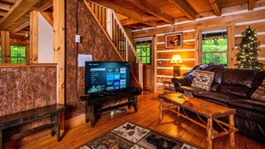 A rustic cabin interior with a wooden staircase, a television displaying streaming services, a leather sofa, and a Christmas tree. - Romantic Log Cabin w/ Hot Tub, Jetted Tub & Wood Stove (Nashville)