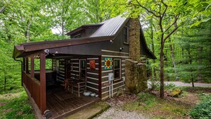 A rustic cabin with a wooden deck and a stone chimney, surrounded by dense forest. - Romantic Log Cabin w/ Hot Tub, Jetted Tub & Wood Stove (Nashville)