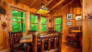A wooden cabin interior with a dining table, chairs, and large windows offering a view of the forest. - Romantic Log Cabin w/ Hot Tub, Jetted Tub & Wood Stove (Nashville)