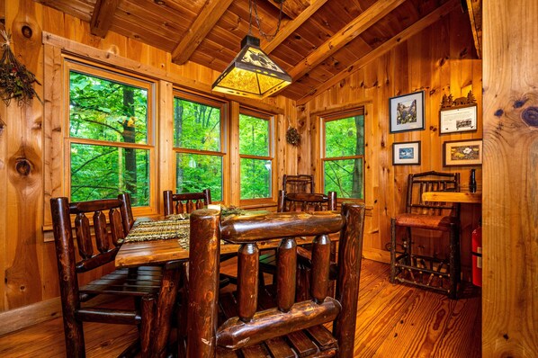 Interior de una cabaña de madera con una mesa de comedor, sillas y ventanas amplias que dan a un bosque.