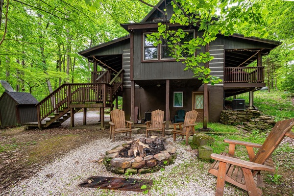 Stone fire pit surrounded by wooden Adirondack chairs sits on gravel beneath the elevated wood cabin deck.