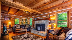 A cozy log cabin interior with a stone fireplace, leather sofas, a Christmas tree, and a television. - Romantic Log Cabin w/ Hot Tub, Jetted Tub & Wood Stove (Nashville)