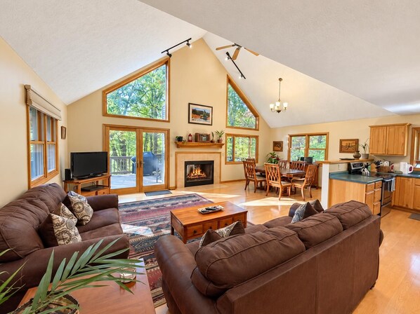 A spacious living area with a fireplace, a dining table, and a kitchen area with wooden cabinets and a large window.