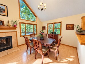 A dining area with a wooden table and chairs, large windows with a view of trees, a fireplace, and a chandelier.