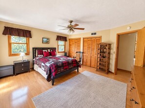 A bedroom with a bed, a nightstand, a chair, a bookshelf, a ceiling fan, and a window with a view of trees.