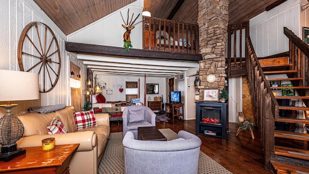 A spacious living room with a high ceiling, a stone fireplace, a wooden staircase, and a cozy seating area with a sofa and armchair.