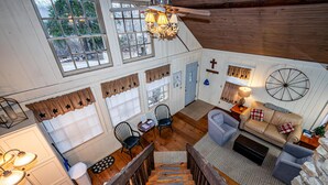 A cozy cabin interior with a wooden staircase, a dining area, a living room with a sofa and armchairs, and a large window overlooking a snowy landscape.