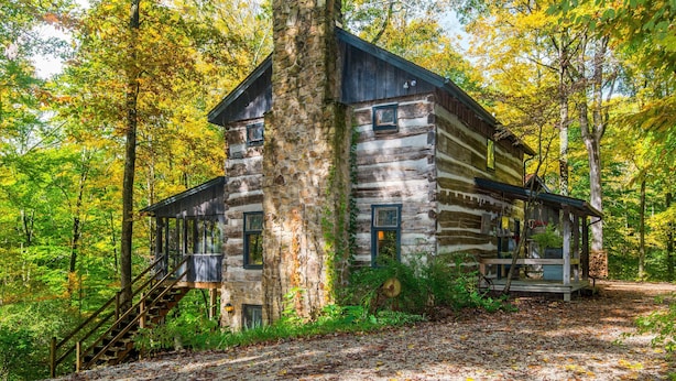Une cabane en rondins avec une cheminée en pierre, une véranda en bois et un escalier menant à l’entrée.