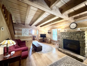Interior de cabana de troncos com lareira de pedra, vigas de madeira, televisão e um sofá vermelho.