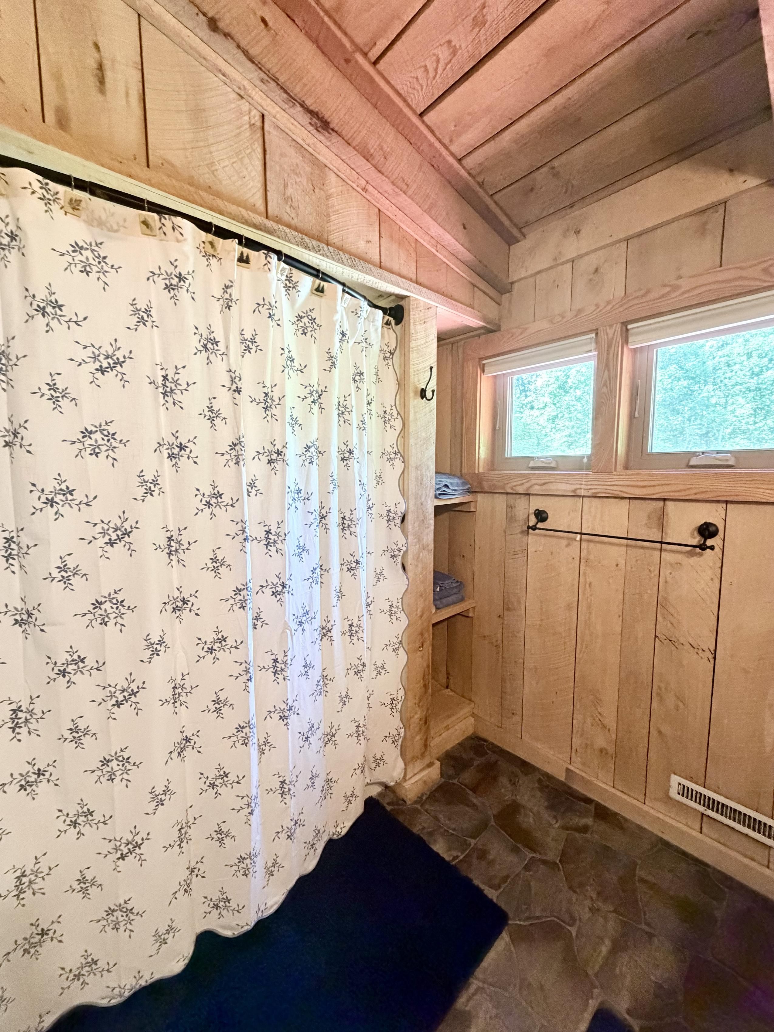 A bathroom with wooden walls, a shower curtain with a floral pattern, a window, and a stone floor.