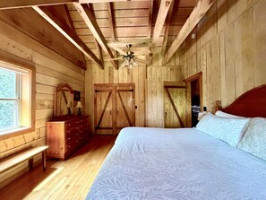 A bedroom with a wooden ceiling, a bed with patterned bedding, a dresser, a mirror, and a window. - Secluded Log Cabin | Hot Tub, Fireplace & Near Nashville 2BR, 1.5BA (Nashville)