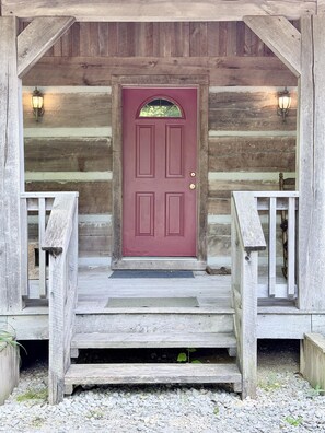 A wooden cabin with a red door, a porch with steps, and two wall-mounted lights.