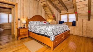 A wooden-paneled bedroom with a sloped ceiling, a wooden bed with a patterned bedspread, a nightstand with a lamp, and a window with a view. - Secluded Log Cabin | Hot Tub, Fireplace & Near Nashville 2BR, 1.5BA (Nashville)