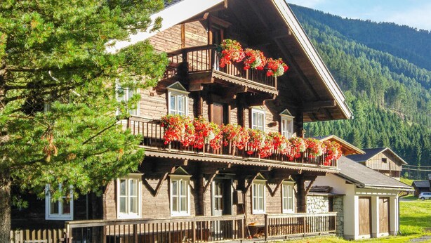 A traditional wooden chalet with a balcony adorned with red flowers, set against a mountainous backdrop.