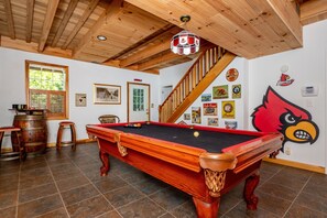 Una mesa de billar en una sala con vigas de madera, una escalera y varias decoraciones en las paredes, incluyendo recuerdos deportivos.