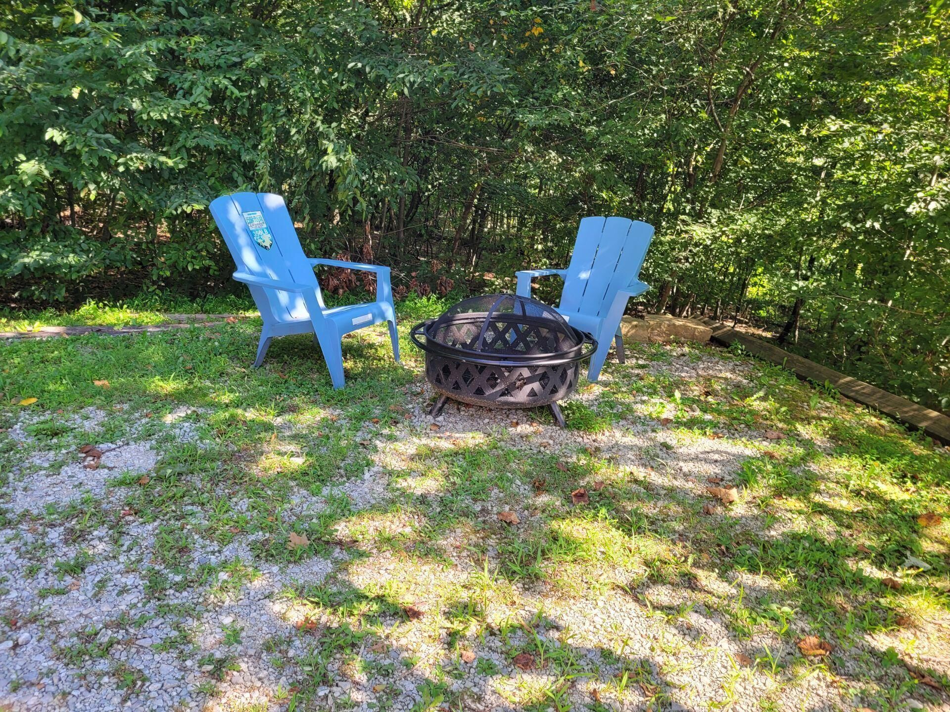 A fire pit with two blue Adirondack chairs in a grassy area surrounded by trees.