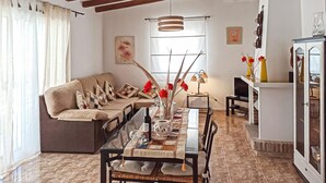 Uma sala de estar aconchegante com sofá, mesa de jantar e cadeiras. Há uma janela com cortinas, um lustre e um armário com objetos decorativos.