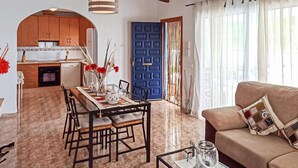 Uma cozinha moderna com armários de madeira, uma mesa de jantar com cadeiras, uma porta azul e uma sala de estar com sofá e mesa de centro.