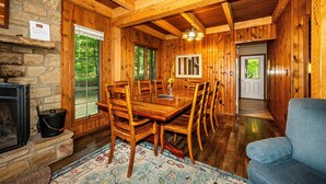 Une salle à manger en bois, avec une grande table et des chaises, une cheminée en pierre et une vue sur l’extérieur grâce à de grandes fenêtres.