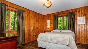 Une chambre aux murs lambrissés, comprenant un lit, un bureau et deux fenêtres avec des rideaux.