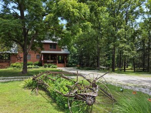 Una baita rustica con veranda, circondata da alberi fitti e una grande ruota arrugginita.