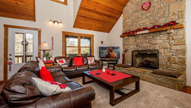 A cozy living room with a stone fireplace, leather sofa, and a television. - Private Lake Lodge | 21 Acres, Hot Tub & Game Room 5BR, 2.5BA (Morgantown)