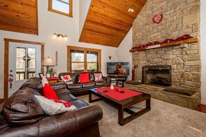 A cozy living room with a stone fireplace, leather sofa, and a television. - Private Lake Lodge | 21 Acres, Hot Tub & Game Room 5BR, 2.5BA (Morgantown)