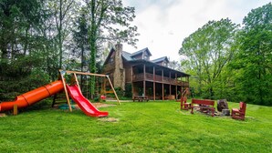 A wooden cabin with a porch, a red slide, and picnic tables in a grassy yard. - Private Lake Lodge | 21 Acres, Hot Tub & Game Room 5BR, 2.5BA (Morgantown)