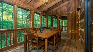 A wooden deck with a dining table and chairs, surrounded by large windows with string lights. - Private 10-Acre Log Cabin | Hot Tub & Near State Park 2BR, 2.5 BA (Nashville)