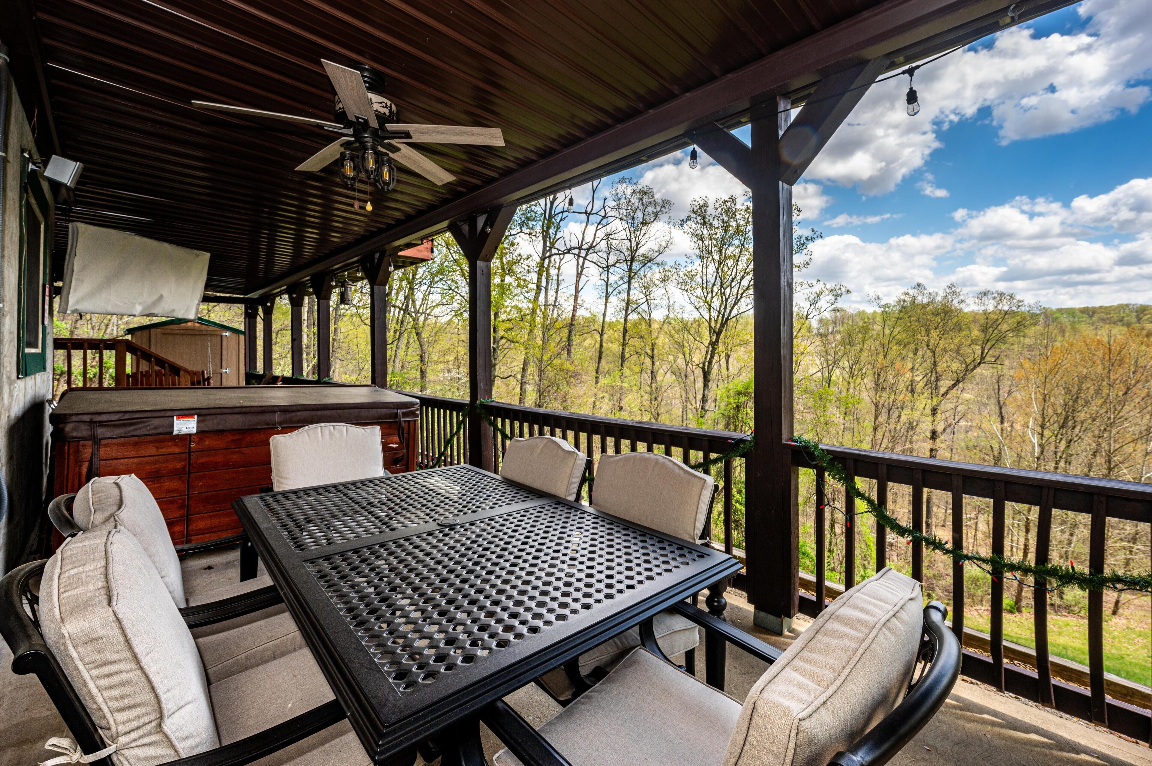 A covered outdoor patio with a table and chairs, a ceiling fan, and a view of trees and hills.