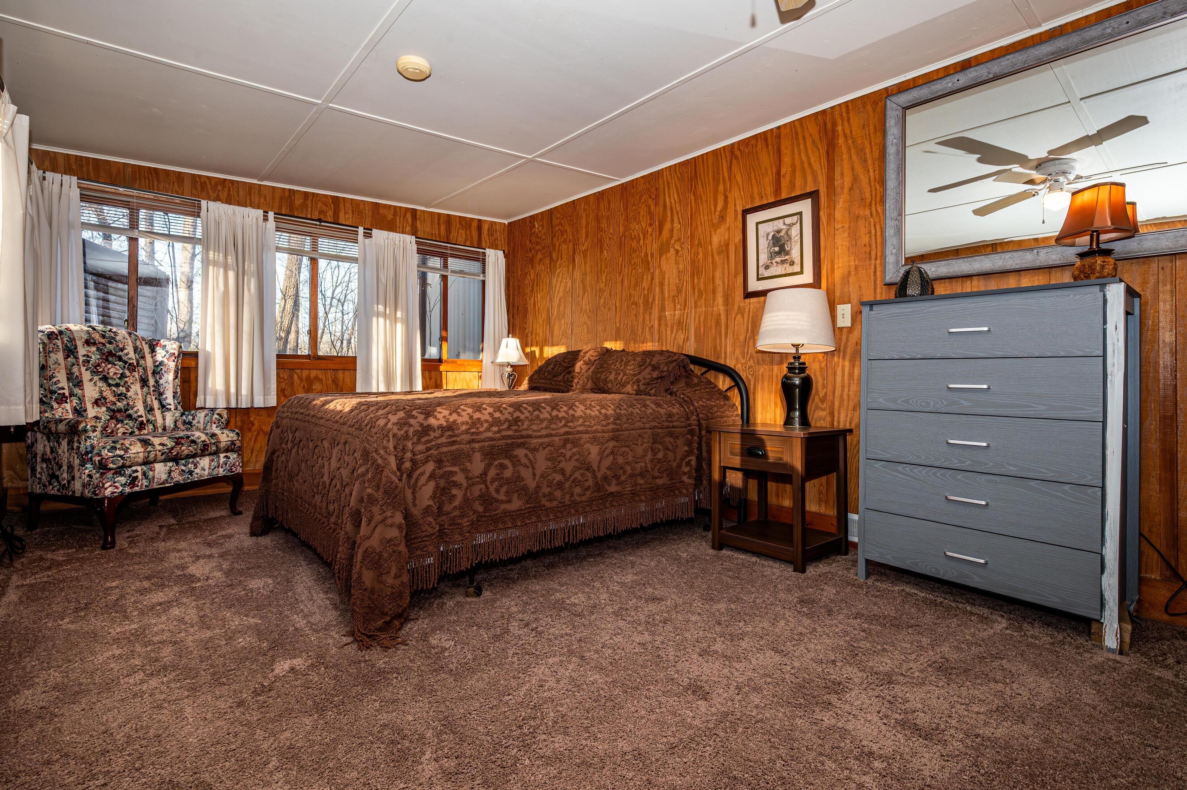 Bedroom features brown patterned bedding, wood-paneled walls, a floral armchair, and a large mirror above a gray dresser.