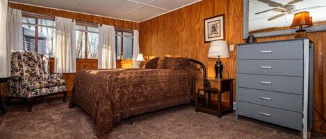 Bedroom features brown patterned bedding, wood-paneled walls, a floral armchair, and a large mirror above a gray dresser.
