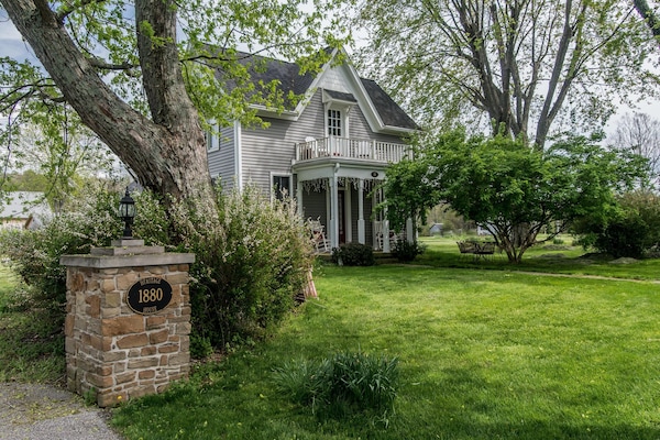 Stone pillar with house number stands beside a gray two-story house with white trim and a second-floor balcony.