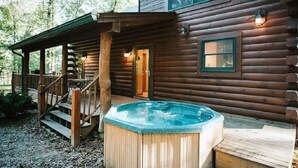 Uma cabana de madeira com banheira de hidromassagem no deck, varanda coberta e escadaria que leva à entrada.