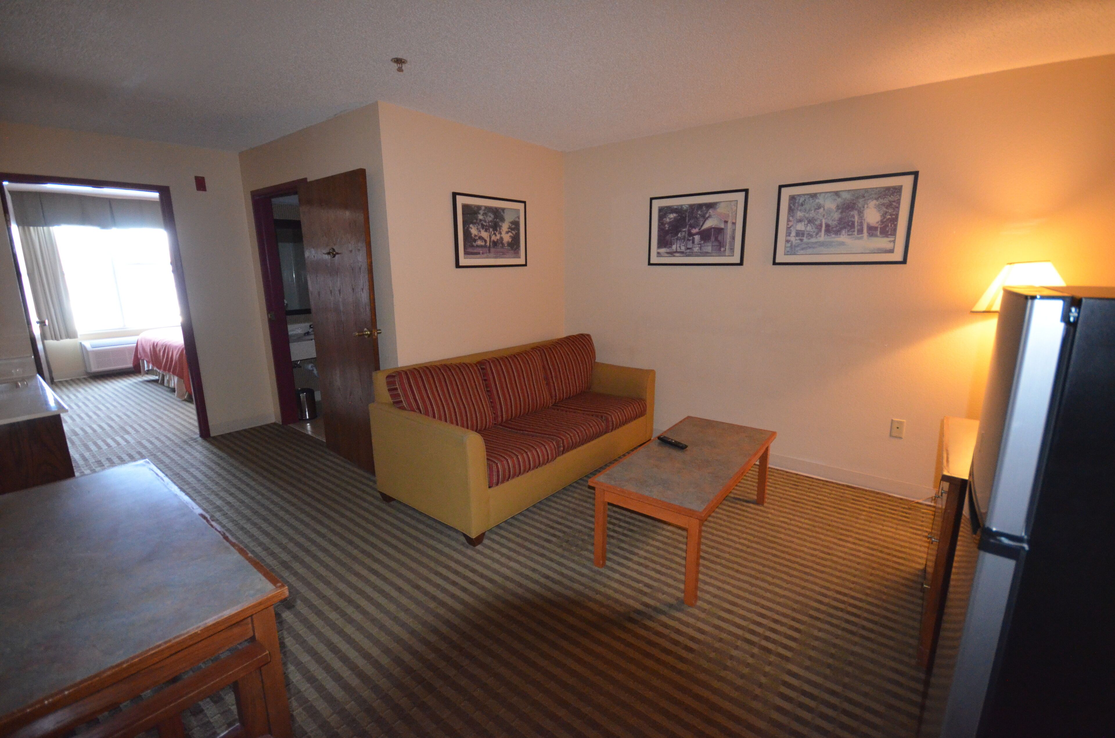 Habitación de hotel con sofá, una mesita, una lámpara y un refrigerador. Hay tres cuadros en la pared.