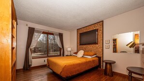 Ein Schlafzimmer mit Holzboden, einem Bett mit brauner Steppdecke, einem Fenster mit Vorhängen, einem Nachttisch, einem Spiegel und einem kleinen hölzernen Hocker.