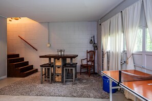A room with a ping pong table, a wooden table with chairs, a staircase, and a window with curtains.