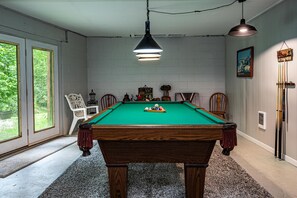 A pool table with a green felt surface, surrounded by chairs, in a room with white brick walls and a large window.