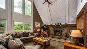 A cozy living room with a stone fireplace, a large window, and a ceiling fan. - Cabin w/ Pond, Hot Tub & Pool Table | Family-Friendly Retreat 3BR, 2BA (Nashville)